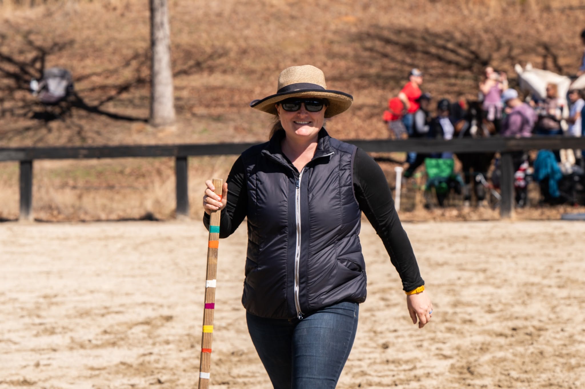 Caroline Templeton in a sun hat carrying a striped course pole, with riders and spectators along the rail behind her
