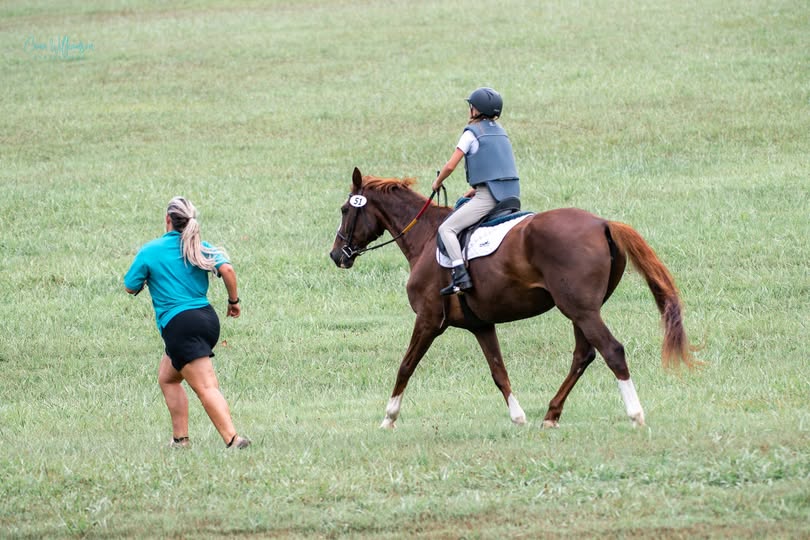 A trainer running alongside a young rider on a chestnut horse across a grassy field
