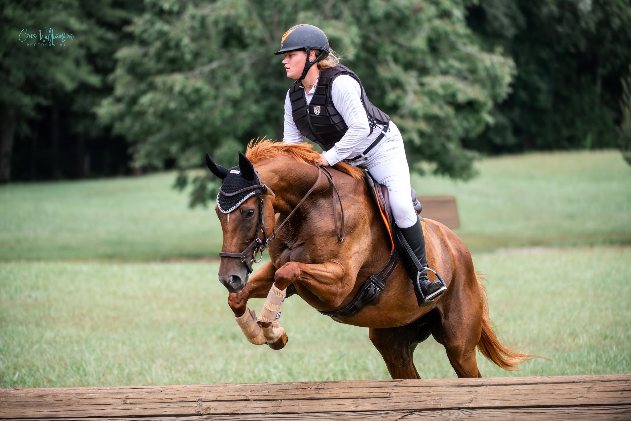 A chestnut horse and rider in cross-country gear jumping a log fence on the grass course